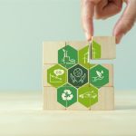 Nine pieces of a cubic wooden puzzle displaying different sustainability logos with a hand placing the last piece on top.