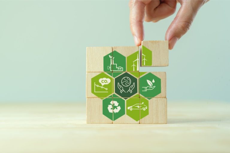 Nine pieces of a cubic wooden puzzle displaying different sustainability logos with a hand placing the last piece on top.