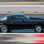 A black car races quickly down an asphalt track. The driver inside is wearing a white helmet and a jacket.