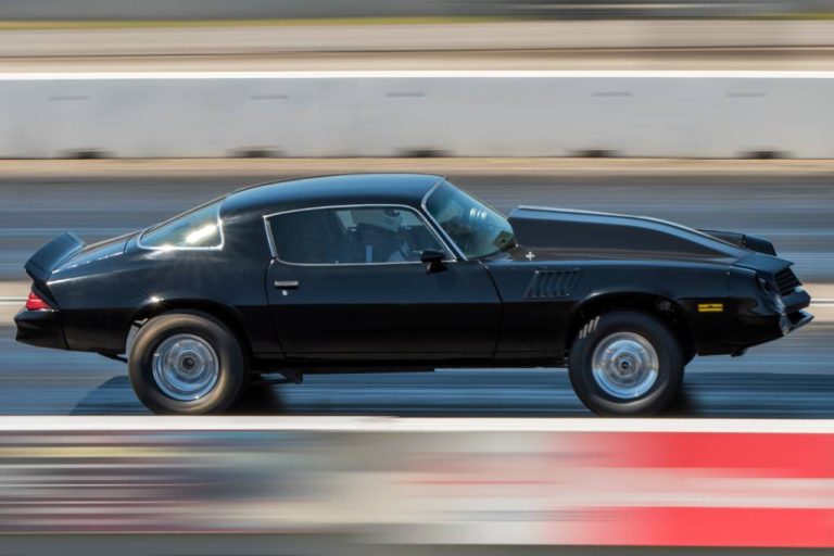 A black car races quickly down an asphalt track. The driver inside is wearing a white helmet and a jacket.
