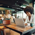 A young woman smiling as she sits at a table in a spacious, cozy coffee shop and types on her laptop.