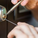 A locksmith is picking a lock on a dark brown door. Each hand is holding a different type of tool.