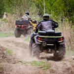 Two ATV riders are driving on a dirt path through the woods. Green trees, shrubs, and dry twigs surround the path.