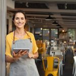 A worker at a restaurant wearing a yellow shirt and gray apron is holding a tablet and greeting customers at the front entrance.