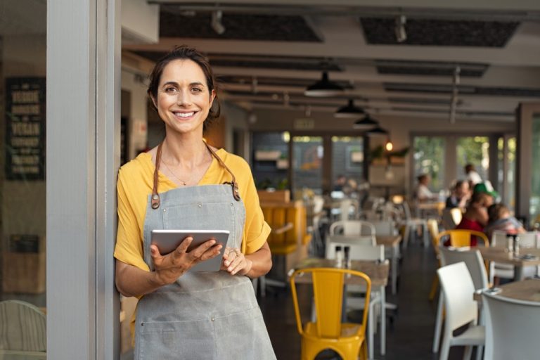 A worker at a restaurant wearing a yellow shirt and gray apron is holding a tablet and greeting customers at the front entrance.