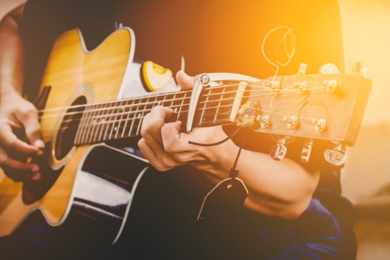 A person wearing a black shirt sitting and playing an acoustic guitar with the sun shining in the background.