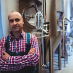 Smiling brewery owner wearing a plaid shirt under an apron. He's standing in front of stainless steel brewery equipment.