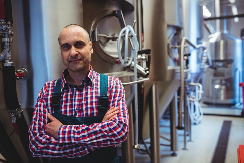 Smiling brewery owner wearing a plaid shirt under an apron. He's standing in front of stainless steel brewery equipment.