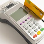 A payment POS terminal sits against a white background with an orange credit card in the machine.