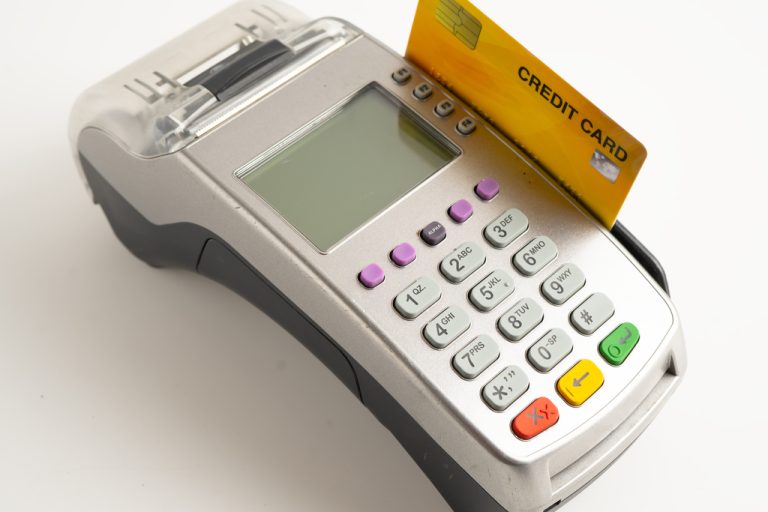 A payment POS terminal sits against a white background with an orange credit card in the machine.