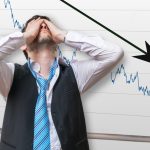 A businessman standing in front of a board with a downward arrow chart. He is covering his face with his hands.