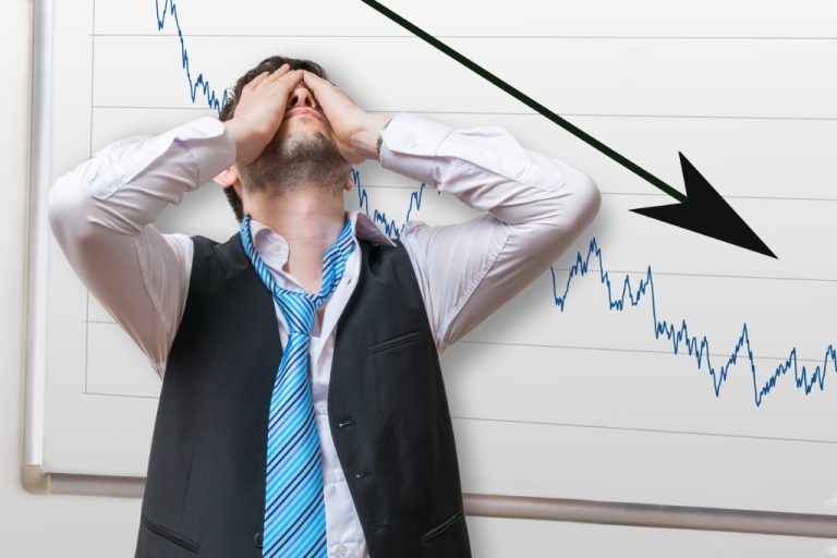 A businessman standing in front of a board with a downward arrow chart. He is covering his face with his hands.