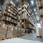 Wide angle view from the ground of stacked products wrapped in plastic on high shelves in various aisles inside a large warehouse.