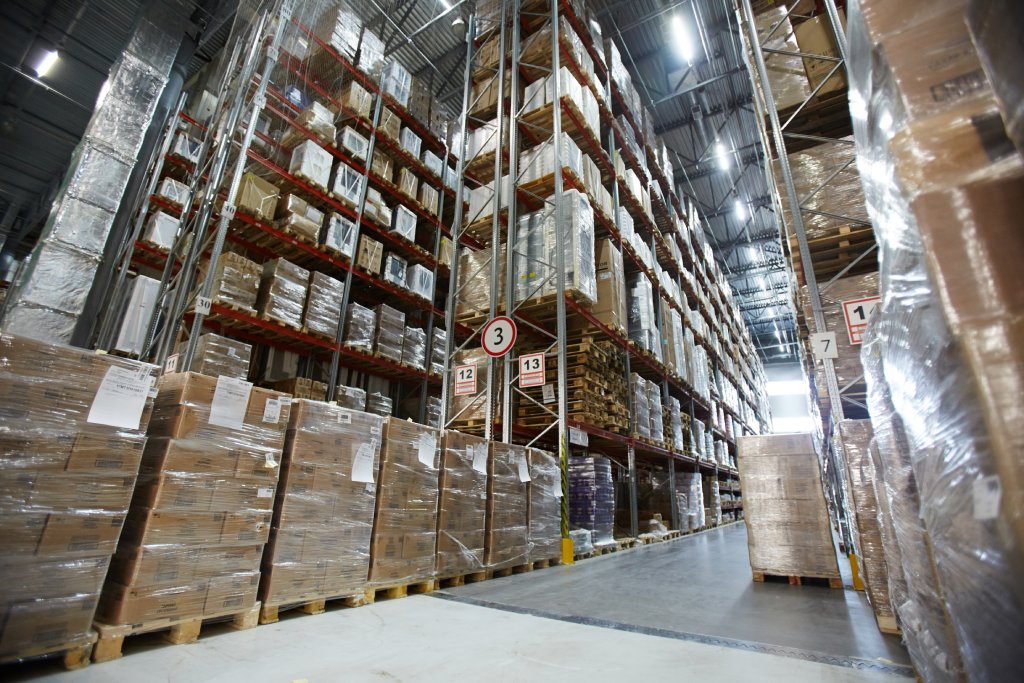 Wide angle view from the ground of stacked products wrapped in plastic on high shelves in various aisles inside a large warehouse.