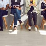 A row of eight, faceless individuals of different genders and races from are waiting in chairs for a job interview.