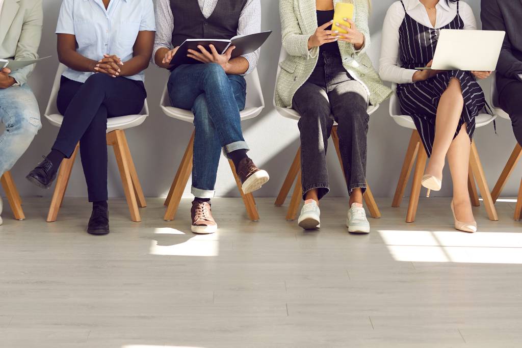 A row of eight, faceless individuals of different genders and races from are waiting in chairs for a job interview.