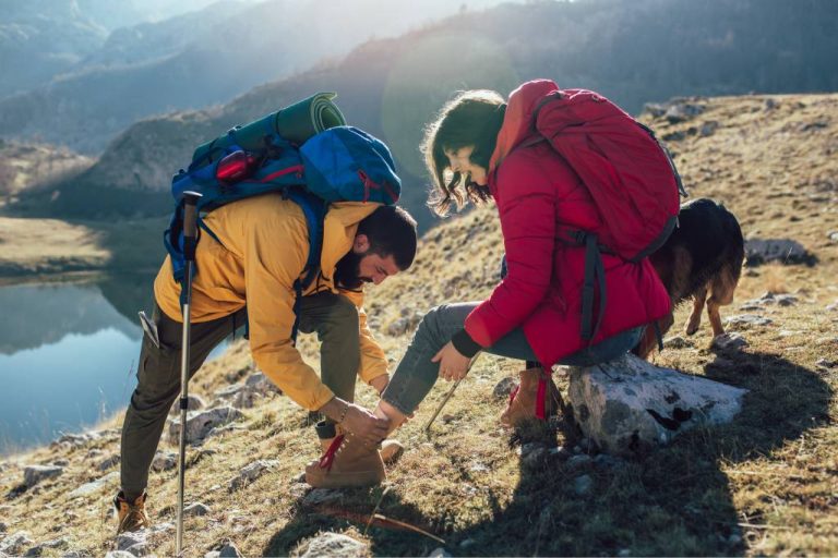 A man in a yellow jacket wraps a bandage on the ankle of a woman wearing a red coat on a hill near a lake.