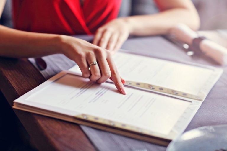 A woman is sitting at a white table and moving her finger down a flat menu. There is wrapped silverware to her right.