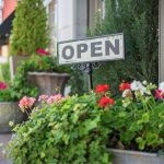 A metal open sign staked into a flowerpot. There are other flowerpots surrounding the exterior of a building.