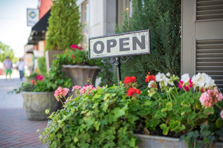 A metal open sign staked into a flowerpot. There are other flowerpots surrounding the exterior of a building.