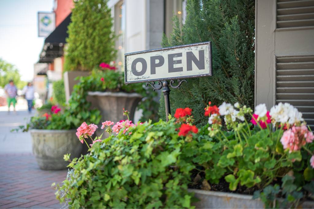 A metal open sign staked into a flowerpot. There are other flowerpots surrounding the exterior of a building.