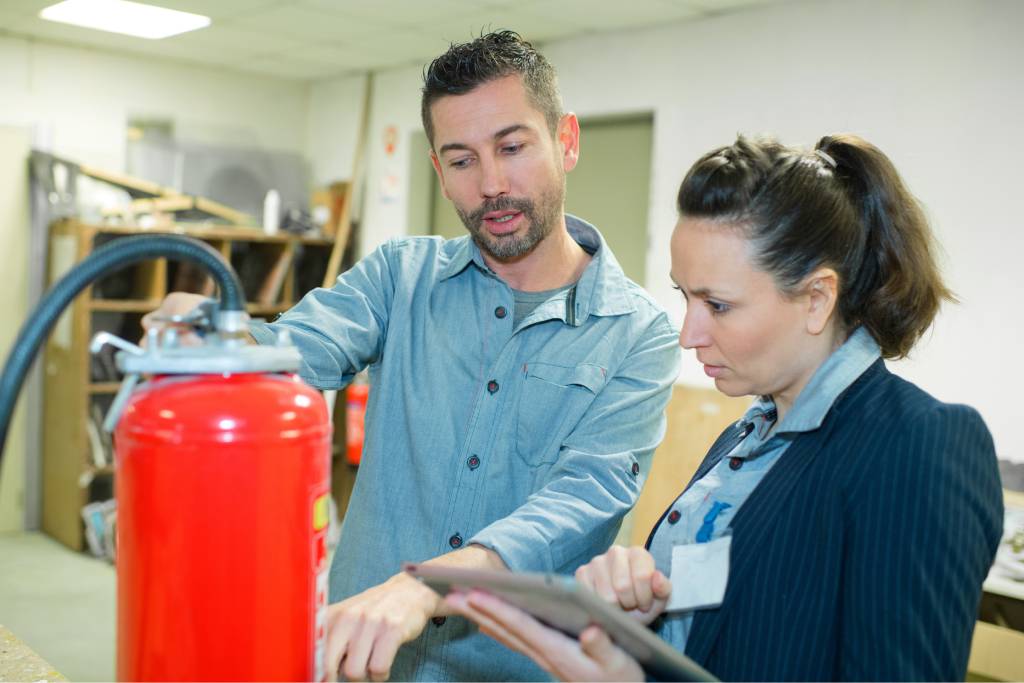 A man and a woman look at a fire extinguisher in a storage room. The man points to the extinguisher while the woman takes notes.