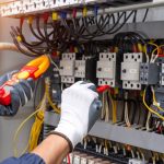 A pair of gloved hands using an AC voltmeter to measure electric current voltage at a circuit breaker.