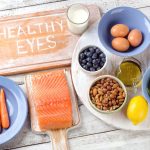 A table containing foods like eggs, salmon, vegetables, and fruits. A wooden cutting board has the words "healthy eyes" on it.