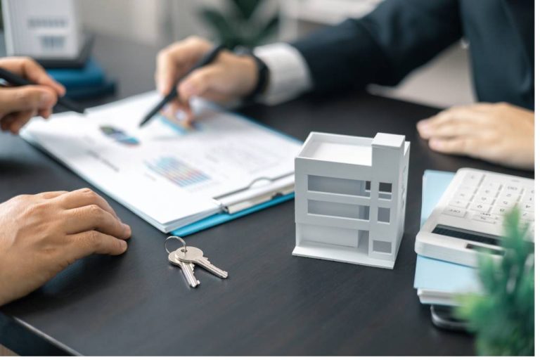A desk with a clipboard, keys, a build model, and two people. One points a pen at the clipboard, and the other rests their hands.