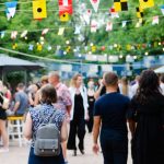 People walk around and sit at booths at an outdoor event. Flags and string lights are hung up above them.