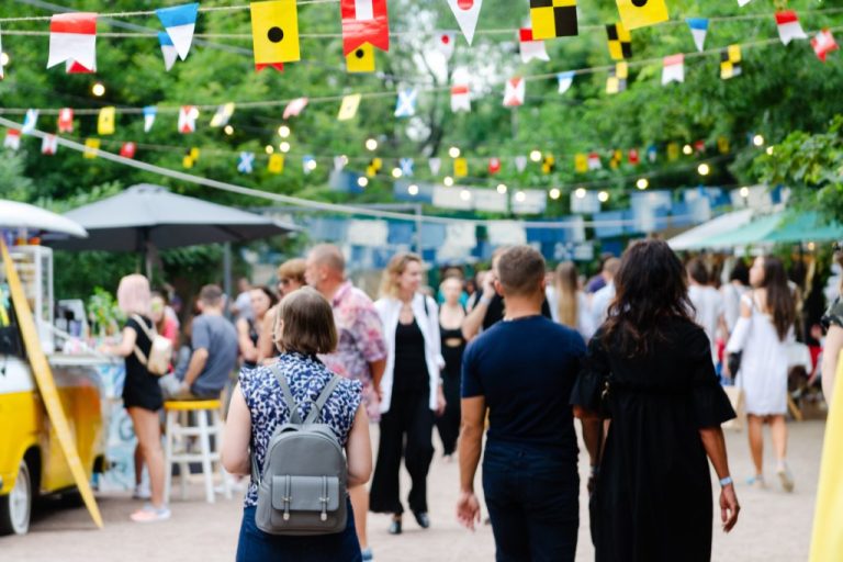 People walk around and sit at booths at an outdoor event. Flags and string lights are hung up above them.