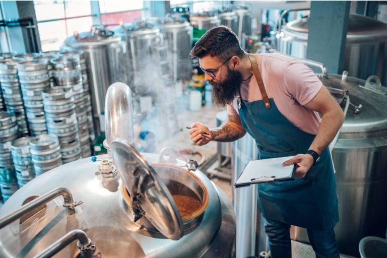 A man with a beard, wearing glasses and an apron, examines a steaming brewing tank in a brewery while holding a clipboard.