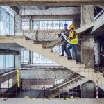 Two employees wearing hard hats are walking up the stairs in a large building that is under construction.
