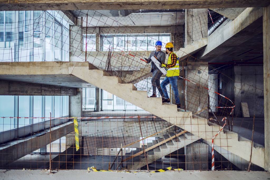 Two employees wearing hard hats are walking up the stairs in a large building that is under construction.