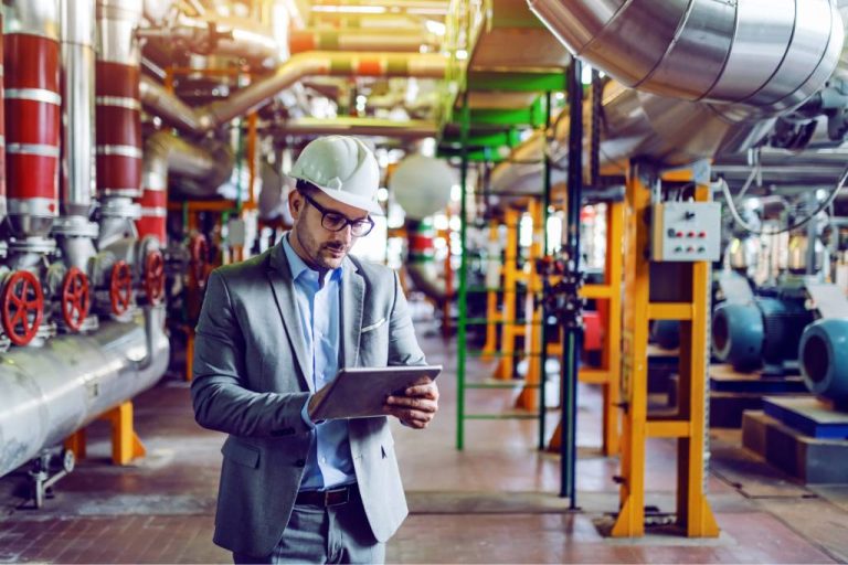 A man in a gray suit and white hard hat looks at a tablet in a production facility.