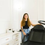A young woman smiling while standing in front of a modern black salon chair in a bright, white room.