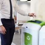 A man wearing a white shirt and a black tie is placing a small plastic water bottle into a recycling bin in an office.