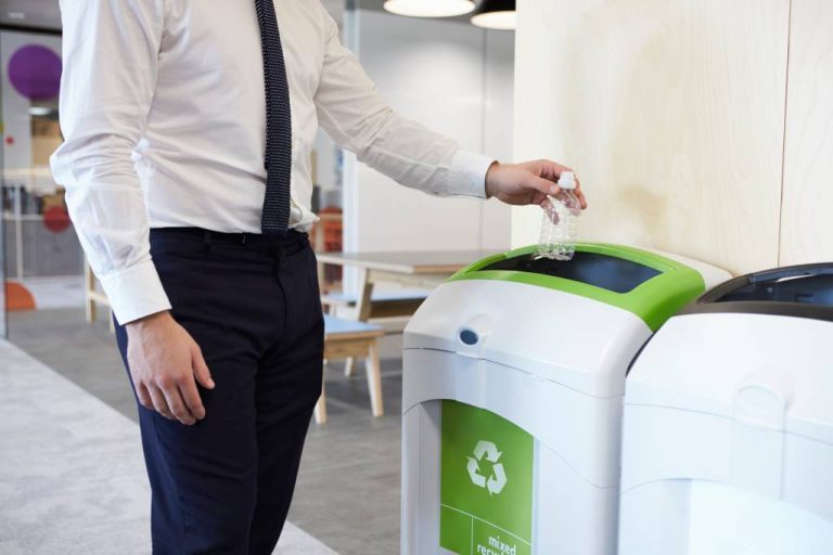 A man wearing a white shirt and a black tie is placing a small plastic water bottle into a recycling bin in an office.