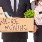 A man and woman stand together wearing business attire. The man holds a box of office supplies reading "we're moving".