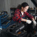 A woman in a blue and red checked shirt with overalls works on the engine of a car. A large blue toolbox is behind her.