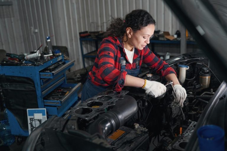 A woman in a blue and red checked shirt with overalls works on the engine of a car. A large blue toolbox is behind her.