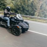A person wearing a black leather jacket, a black helmet, and jeans rides a black trike motorcycle on a road.