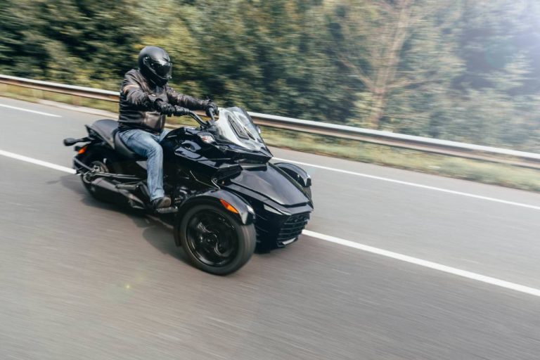 A person wearing a black leather jacket, a black helmet, and jeans rides a black trike motorcycle on a road.