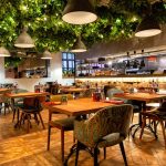 Mismatched chairs placed around small wooden tables in a restaurant's dining room. Greenery covers the ceiling.