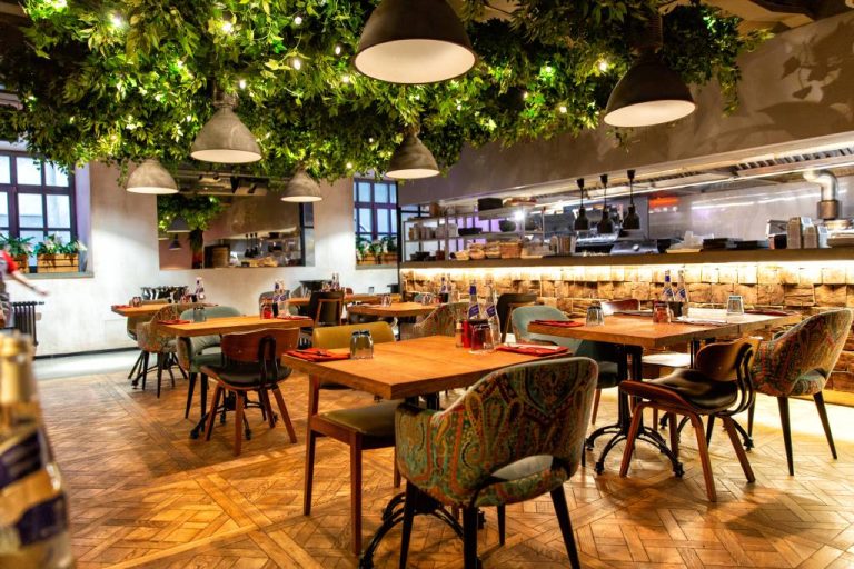 Mismatched chairs placed around small wooden tables in a restaurant's dining room. Greenery covers the ceiling.