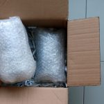 A hand places a product wrapped in bubble wrap into a cardboard box filled with shredded paper and another wrapped product.