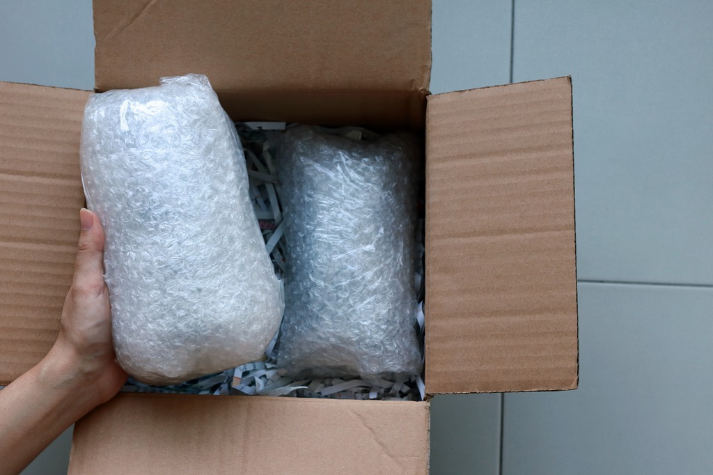 A hand places a product wrapped in bubble wrap into a cardboard box filled with shredded paper and another wrapped product.