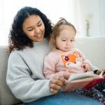 A young woman in a gray sweater reads a picture book to a toddler in a pink sweater sitting on her lap.