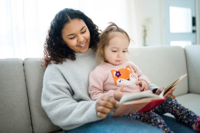 A young woman in a gray sweater reads a picture book to a toddler in a pink sweater sitting on her lap.