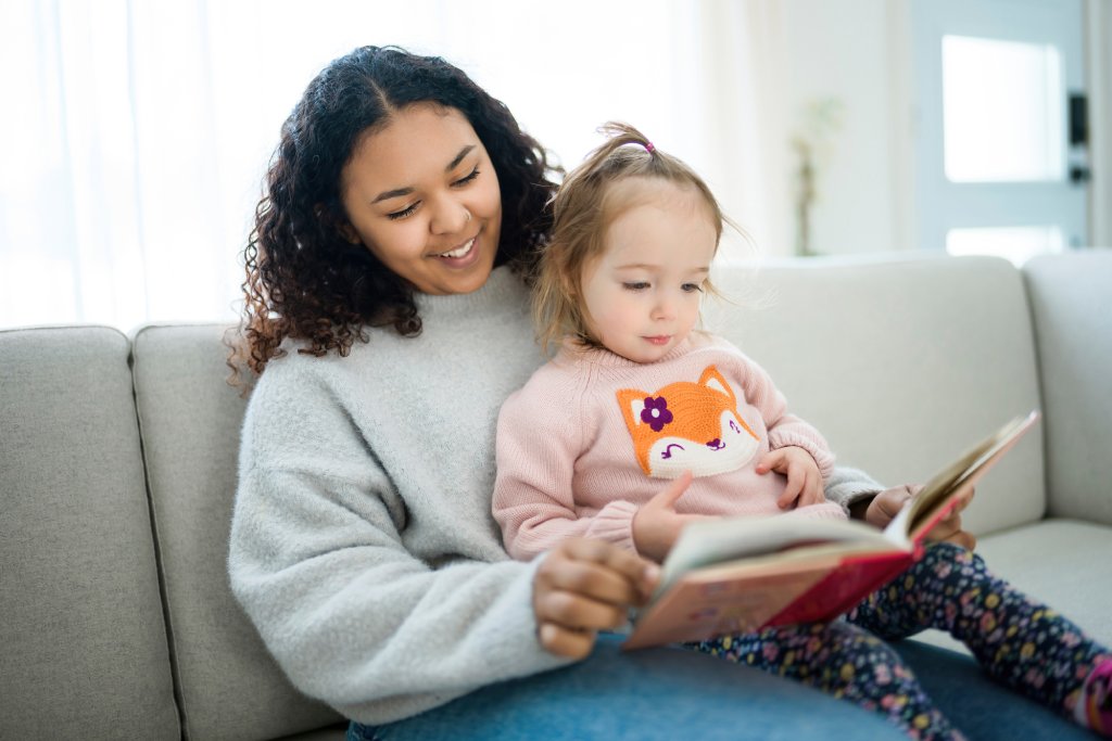 A young woman in a gray sweater reads a picture book to a toddler in a pink sweater sitting on her lap.